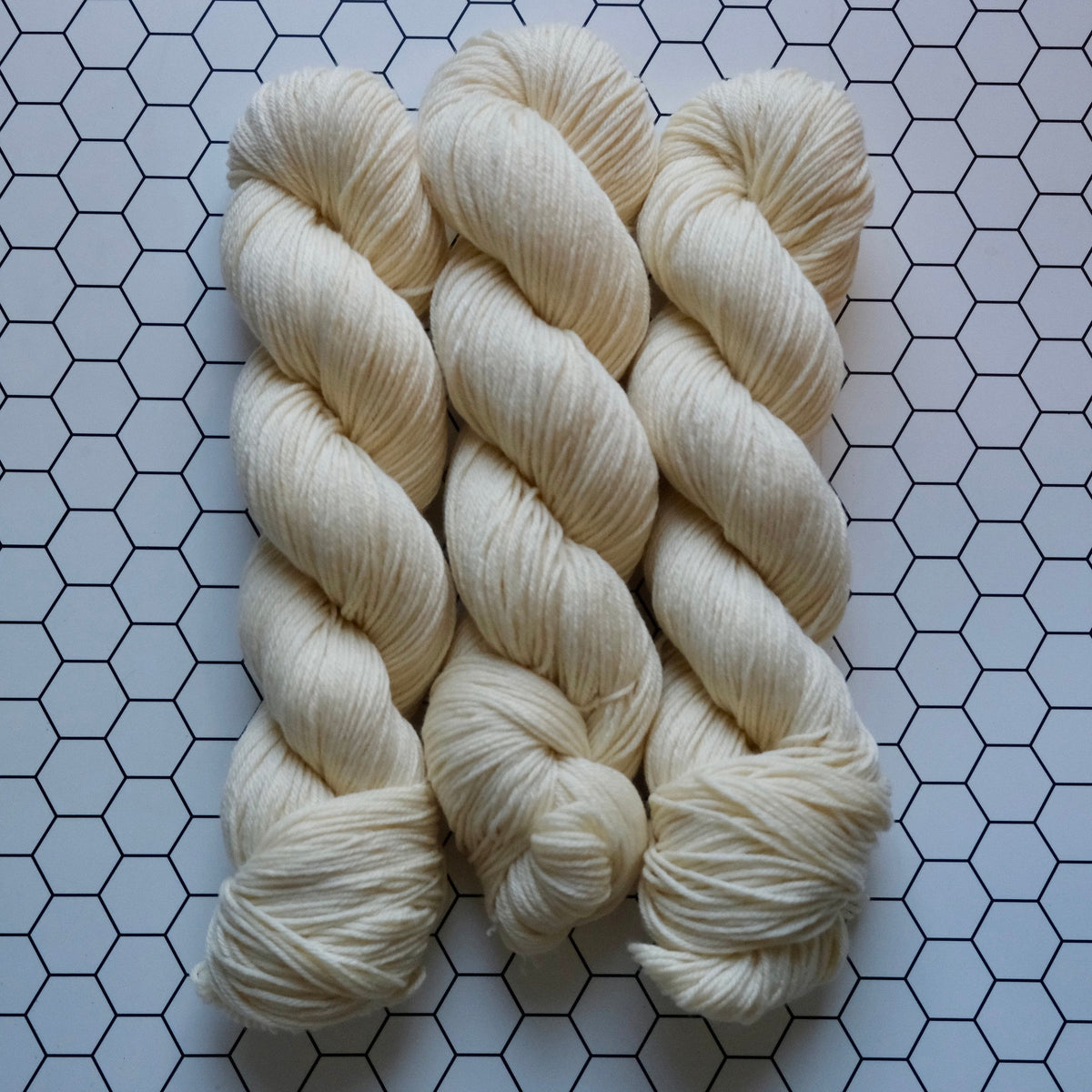 Three skeins of cream-colored yarn arranged vertically on a white surface with a black hexagonal grid pattern.