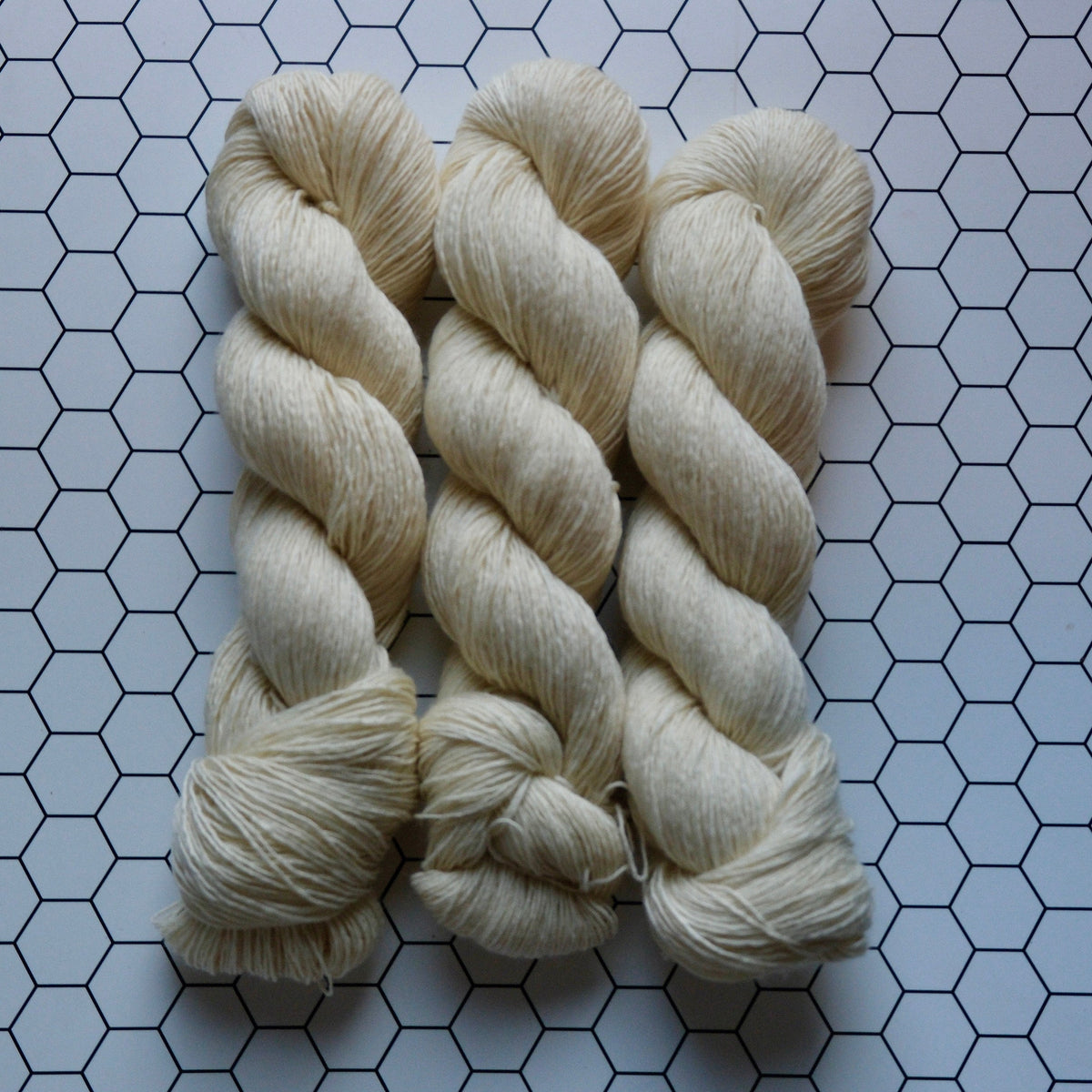 Three skeins of cream-colored yarn arranged vertically on a white surface with a black hexagonal grid pattern.