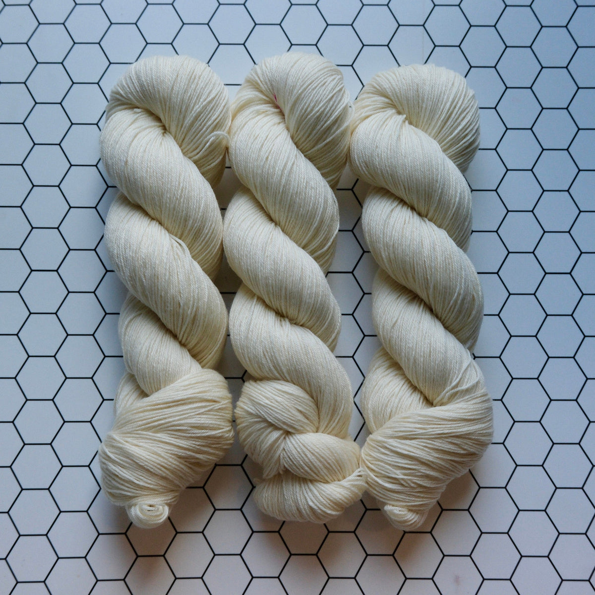 Three skeins of cream-colored yarn arranged vertically on a white surface with a black hexagonal grid pattern.